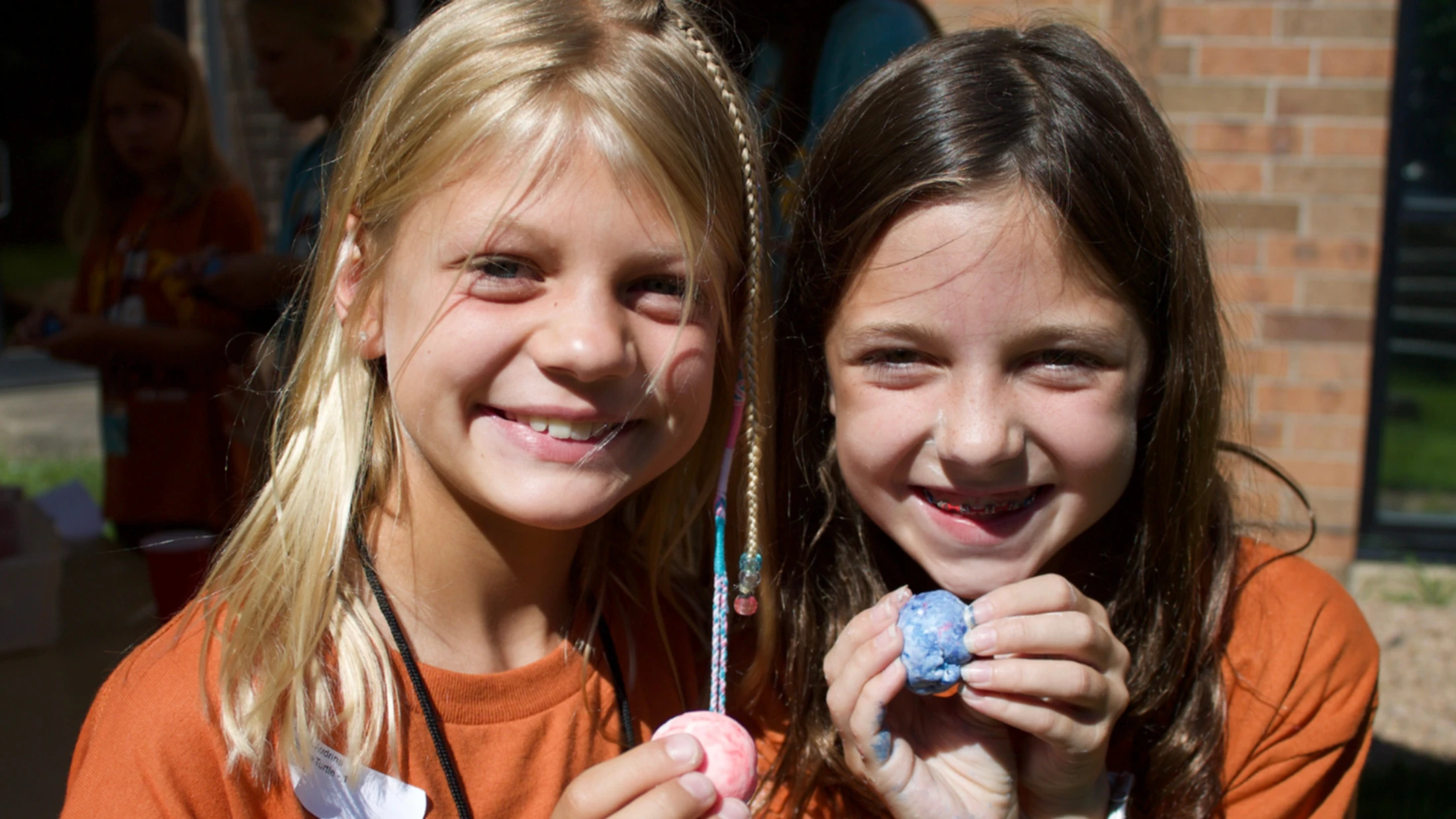 Two smiling young girls in orange shirts proudly hold up small, colorful balls of clay they made during an outdoor activity.