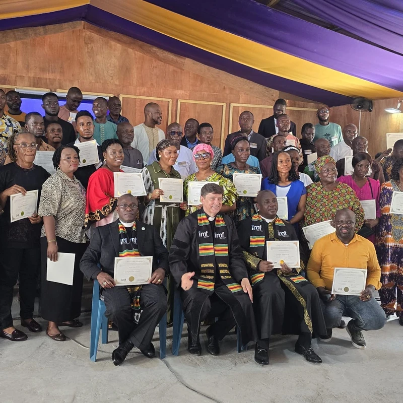 A large, diverse group of men and women pose for a photo, holding up certificates after an International Ministerial Fellowship graduation ceremony.
