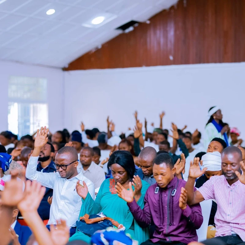 A large congregation of Black men and women sitting indoors with their hands raised in prayer and worship. Many have their eyes closed in deep concentration.