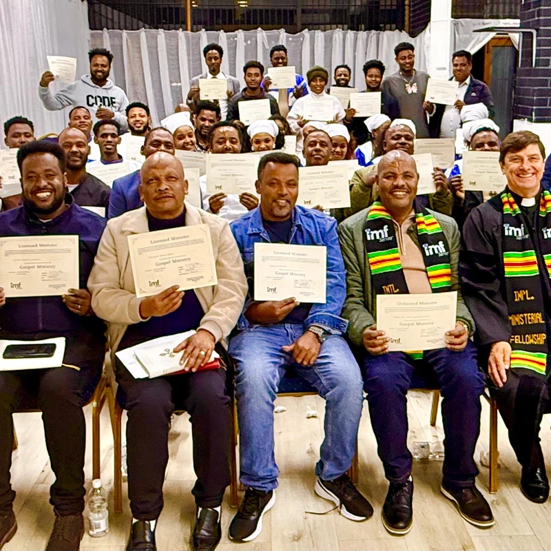 A large, smiling group of men and women pose for a photo, holding up certificates from the IMF Gospel Ministry after a graduation ceremony.