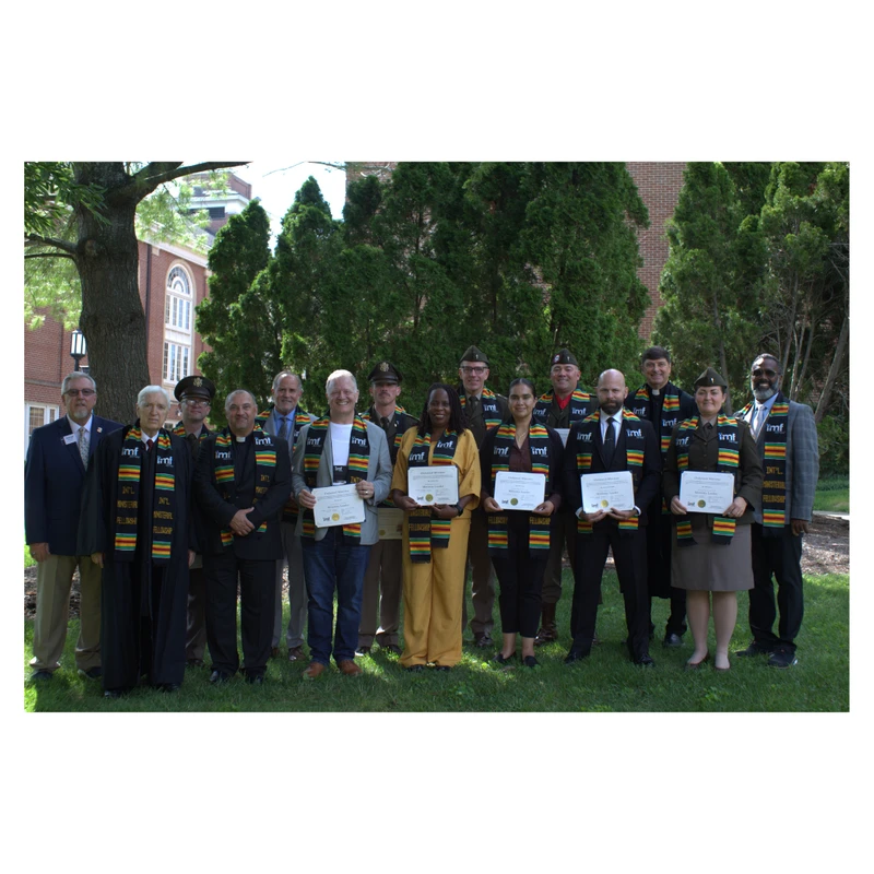 A diverse group of graduates wearing fellowship stoles pose outdoors on a sunny day, holding their certificates in front of a brick building and trees.