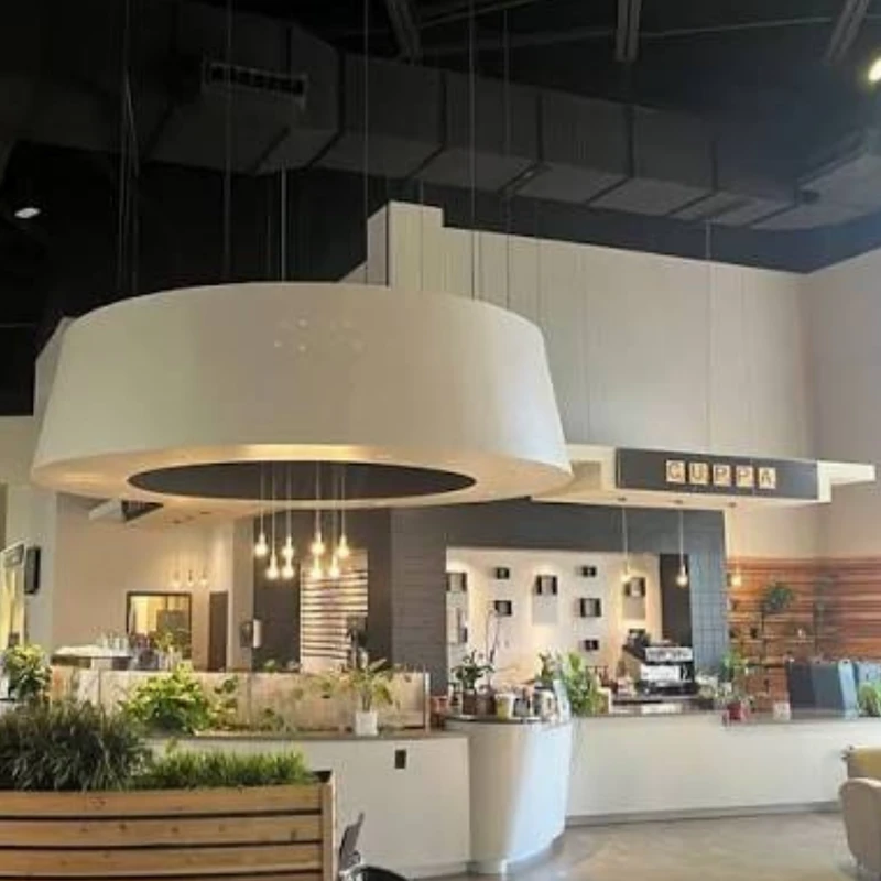 The interior of a modern coffee shop with a large, white circular light fixture hanging from the ceiling over a counter area decorated with green plants.