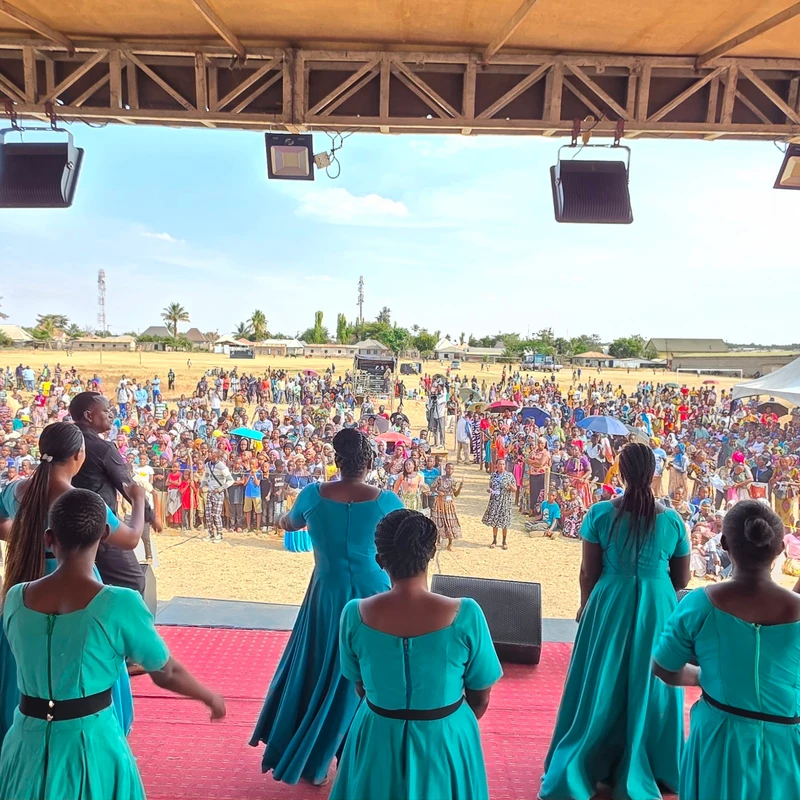 View from behind a choir in turquoise dresses as they perform on a red stage for a large crowd gathered in a sunny field.
