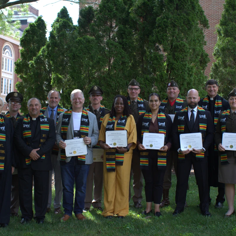 A diverse group of fourteen people, some in uniform, pose on a lawn wearing sashes and holding certificates from the International Ministerial Fellowship.