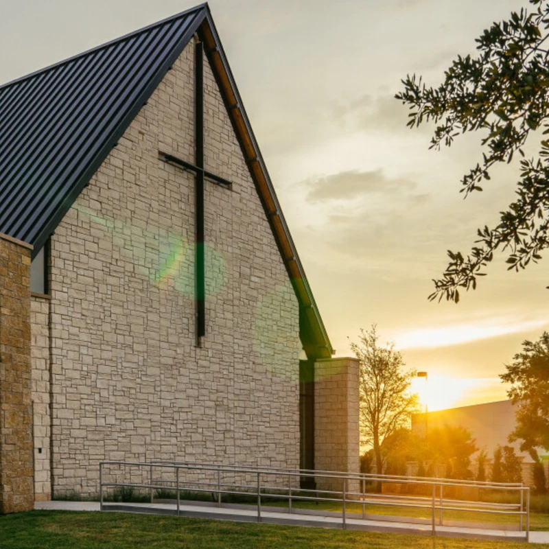 Exterior of a modern stone church with a dark metal roof and large cross on the wall. The sun is setting, casting a warm glow over the scene.