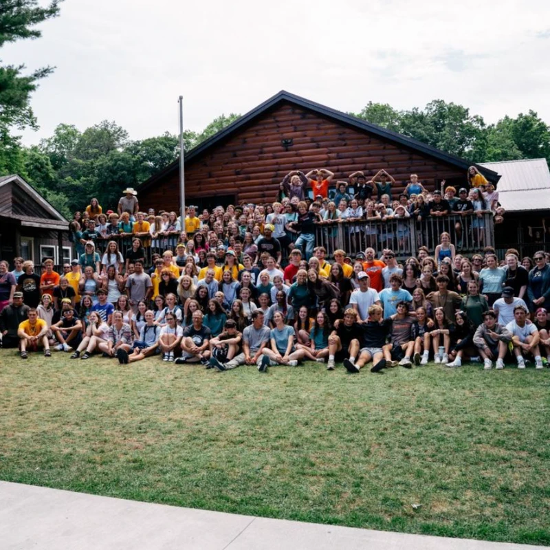 A wide shot of a large group of young people and adults posing for a photo on a grassy lawn in front of a rustic wooden lodge.