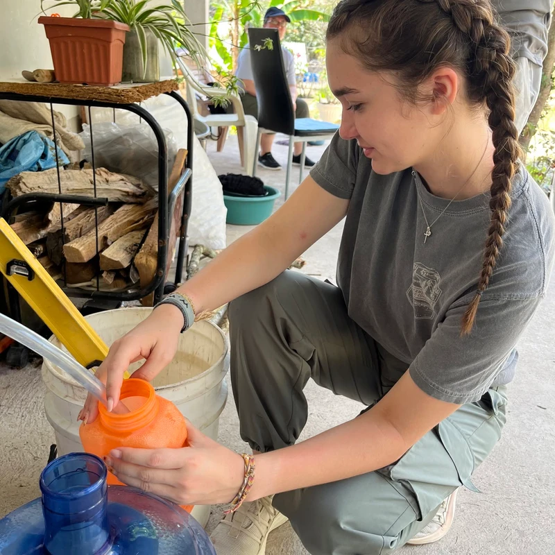 A young woman with braided brown hair crouches on a concrete patio, filling an orange bottle from a clear tube connected to a large water container.