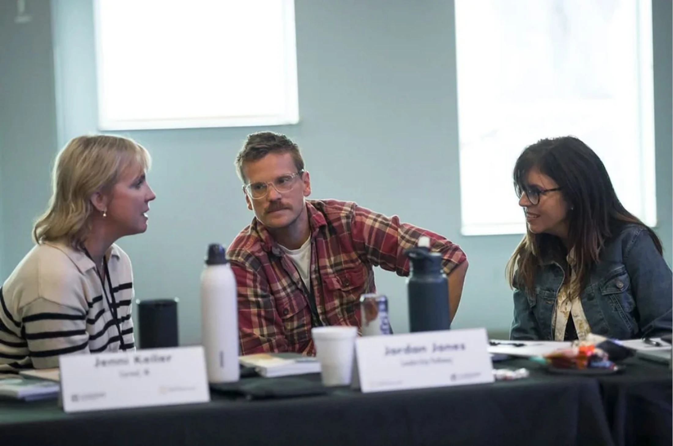 A man and two women sit at a table with name placards, engaged in conversation during what appears to be a conference or workshop.