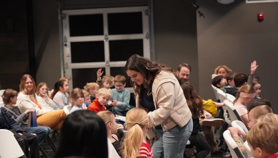 A smiling woman leans over to talk to a young girl in a room full of children sitting on white chairs, with some kids raising their hands in the background.