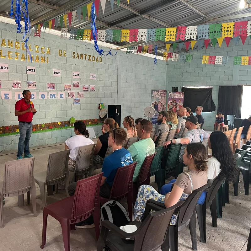 A man in a red shirt speaks with a microphone to an audience seated on plastic chairs in a hall decorated with colorful banners and photos.