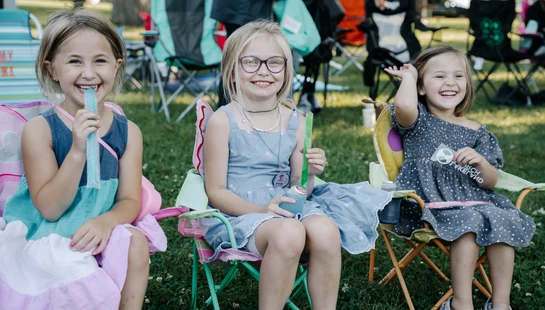 Three young girls sit in colorful camp chairs on the grass, smiling for the camera while eating freeze pops during an outdoor event.