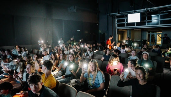 A large group of teenagers sit in a dark auditorium, holding up their smartphones with the flashlights on, creating dozens of small, bright lights.
