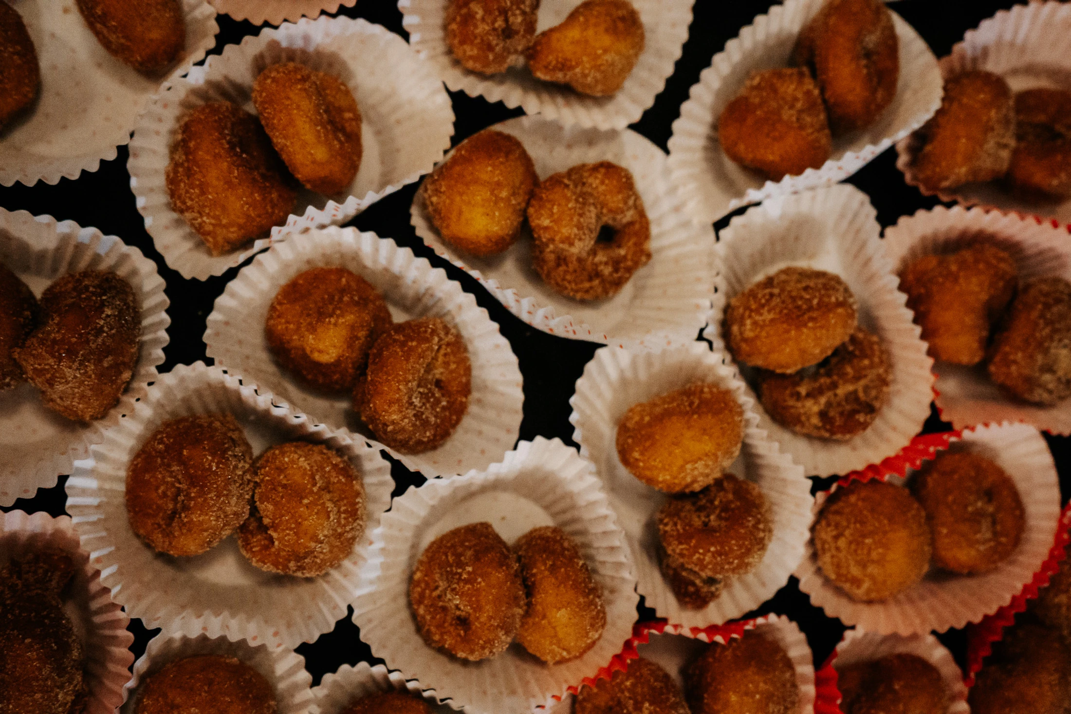 A top-down close-up of many small, golden-brown donuts coated in sugar. They are arranged in pairs inside white, fluted paper liners on a dark surface.