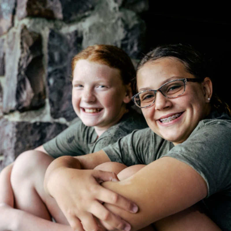 Two young girls in matching green shirts sit side-by-side and smile for the camera. One has red hair and the other wears glasses and braces.