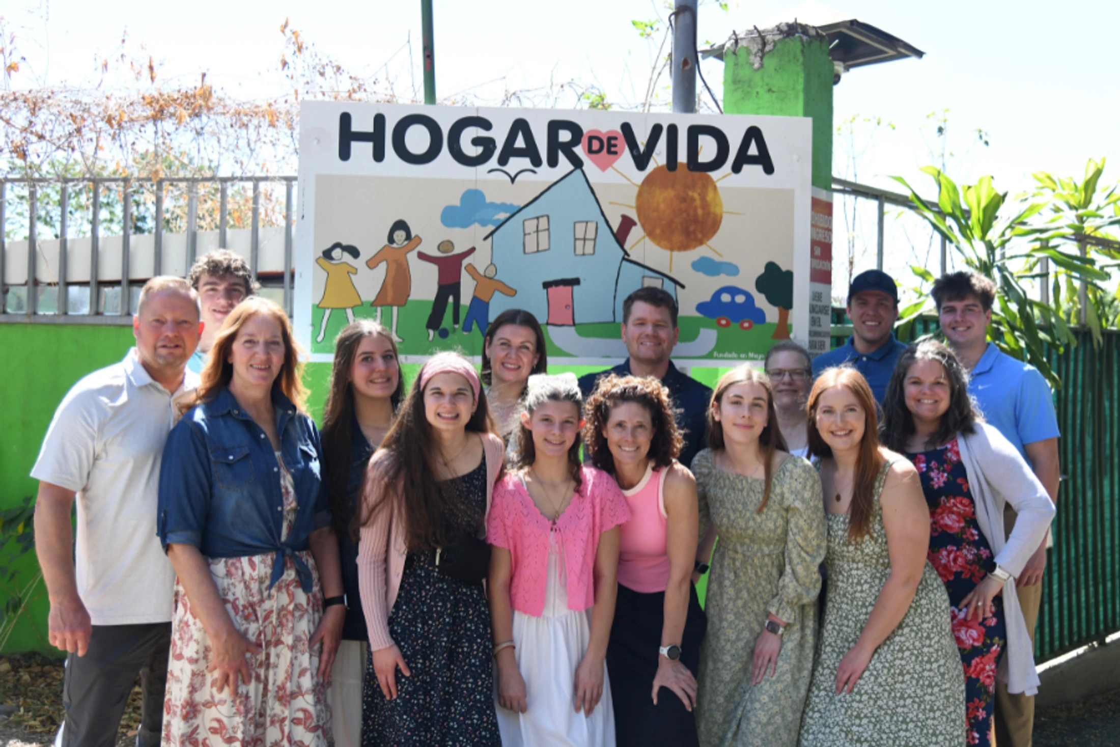 A large group of smiling adults and teenagers stand together outdoors for a portrait in front of a colorful sign that reads 'Hogar de Vida'.