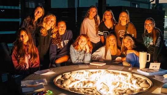 A group of smiling young women are gathered around a modern gas fire pit on a patio at night, looking at the camera for a group photo.