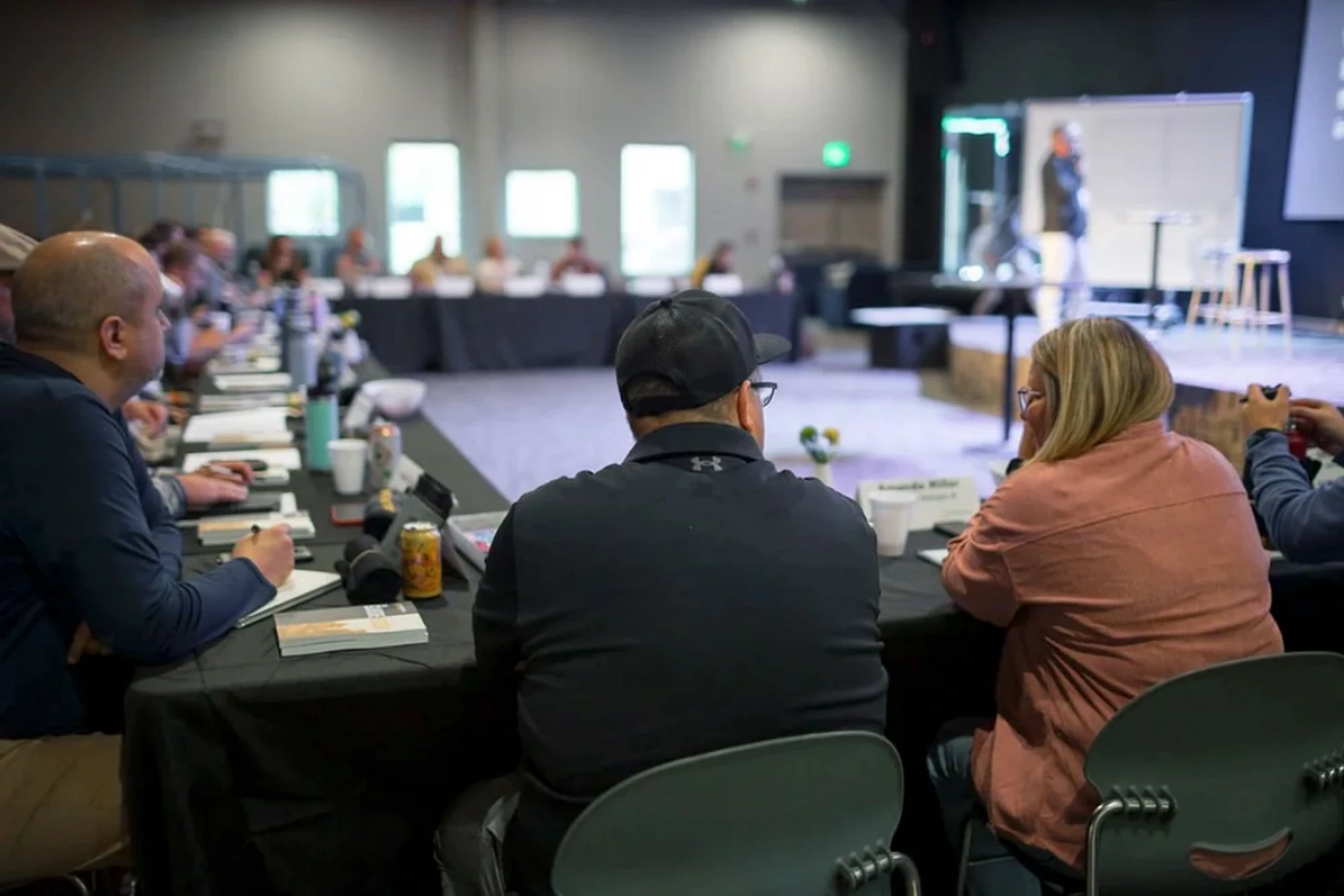 A view from behind attendees sitting at a long table listening to a speaker on a distant stage during a conference or workshop.