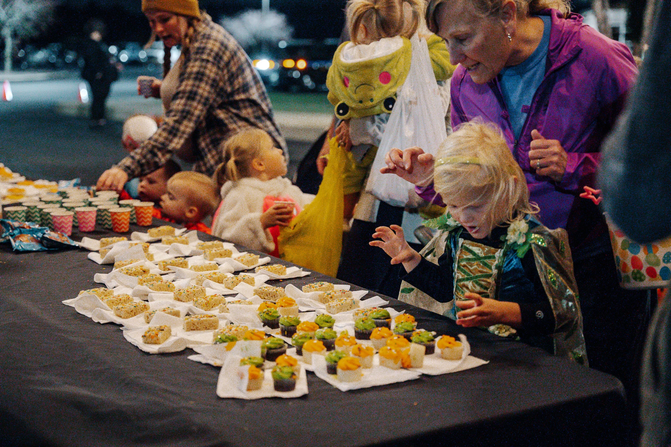 A young girl in a costume excitedly reaches for a cupcake from a table full of treats, surrounded by other children and adults at a nighttime party.