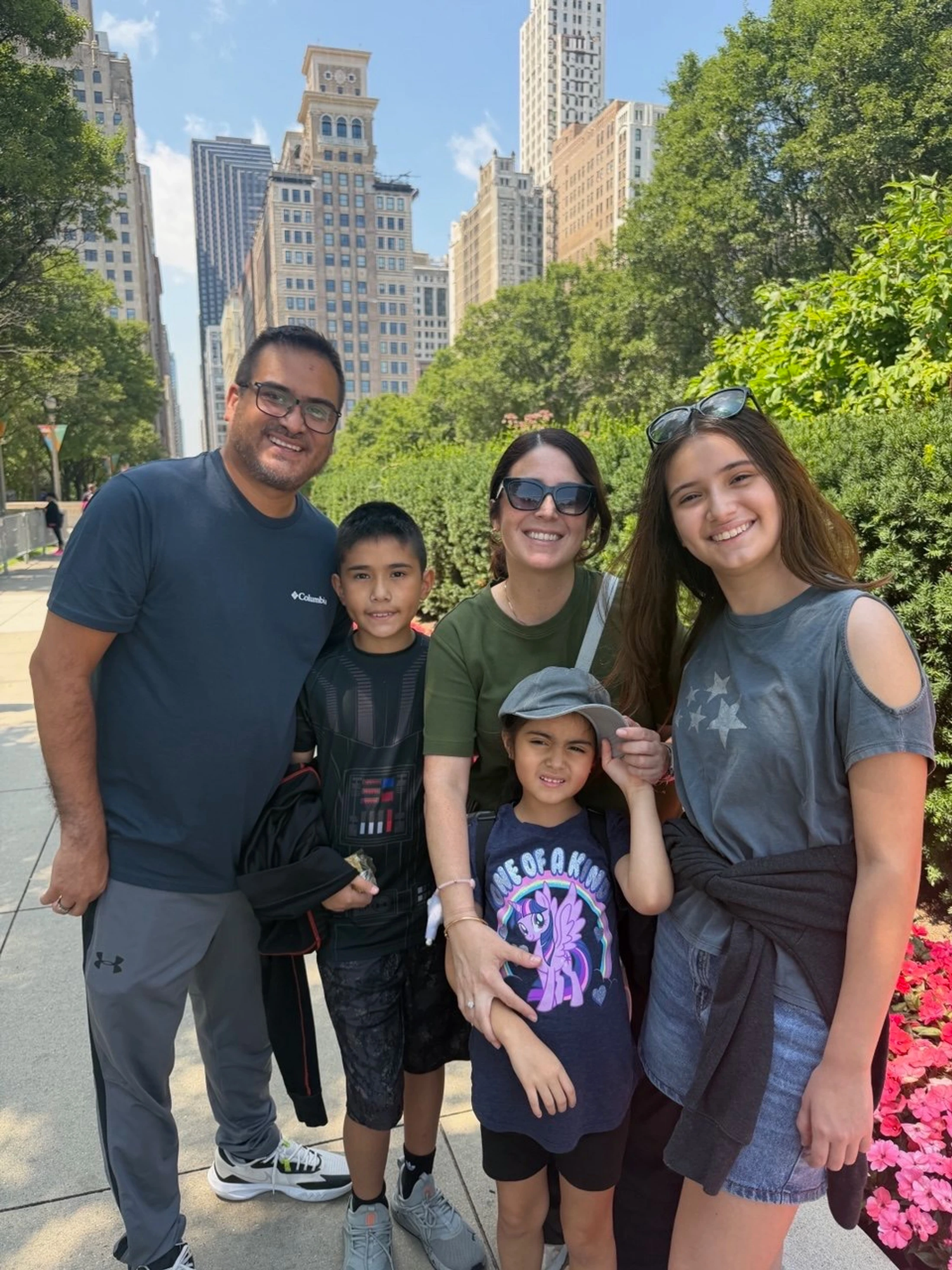 A family of five smile together on a city sidewalk. Parents and three children pose in front of tall buildings and green trees on a sunny day.