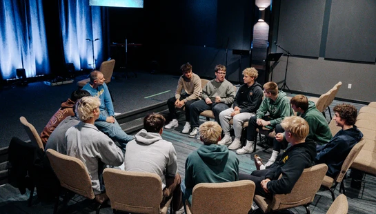 A group of teenage boys and an adult leader sit in a circle of chairs, having a discussion in a large room with a stage in the background.