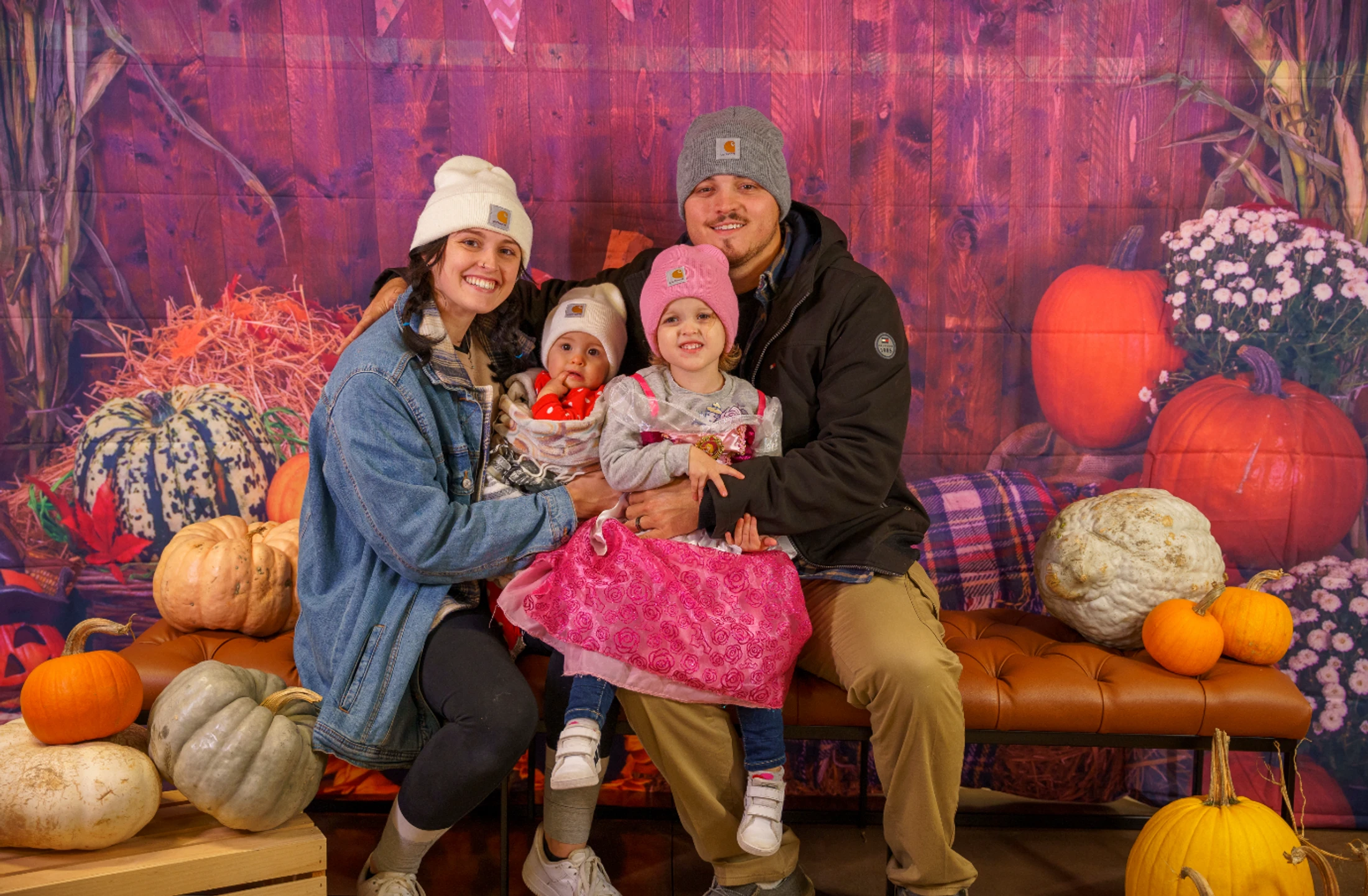 A smiling family of four, a mother, father, young daughter, and baby, pose for a photo in a rustic setting with pumpkins, hay, and a purple wood background.