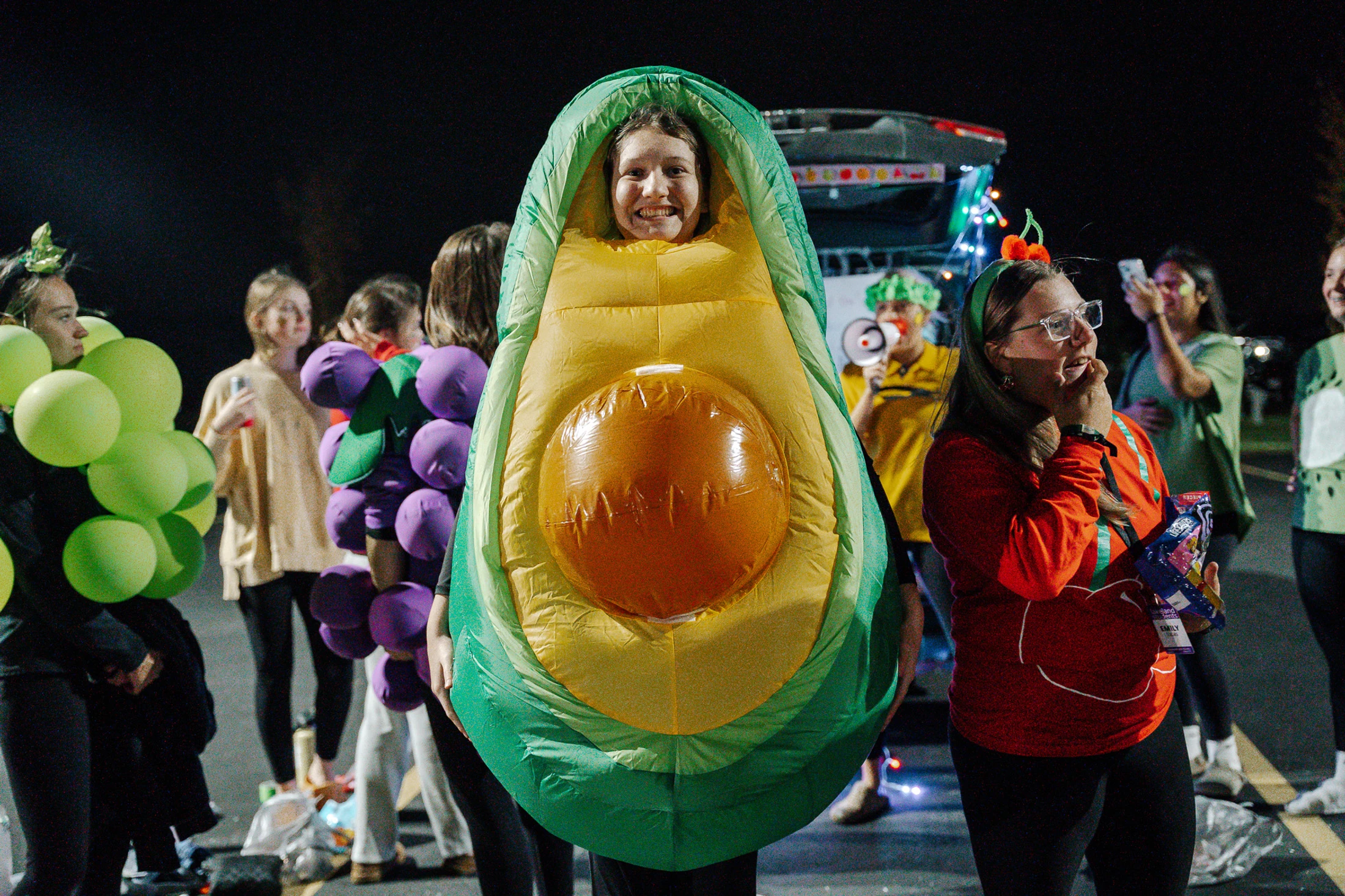 A smiling person wears a large inflatable avocado costume at a nighttime event, surrounded by other people in fruit-themed costumes like grapes and cherries.