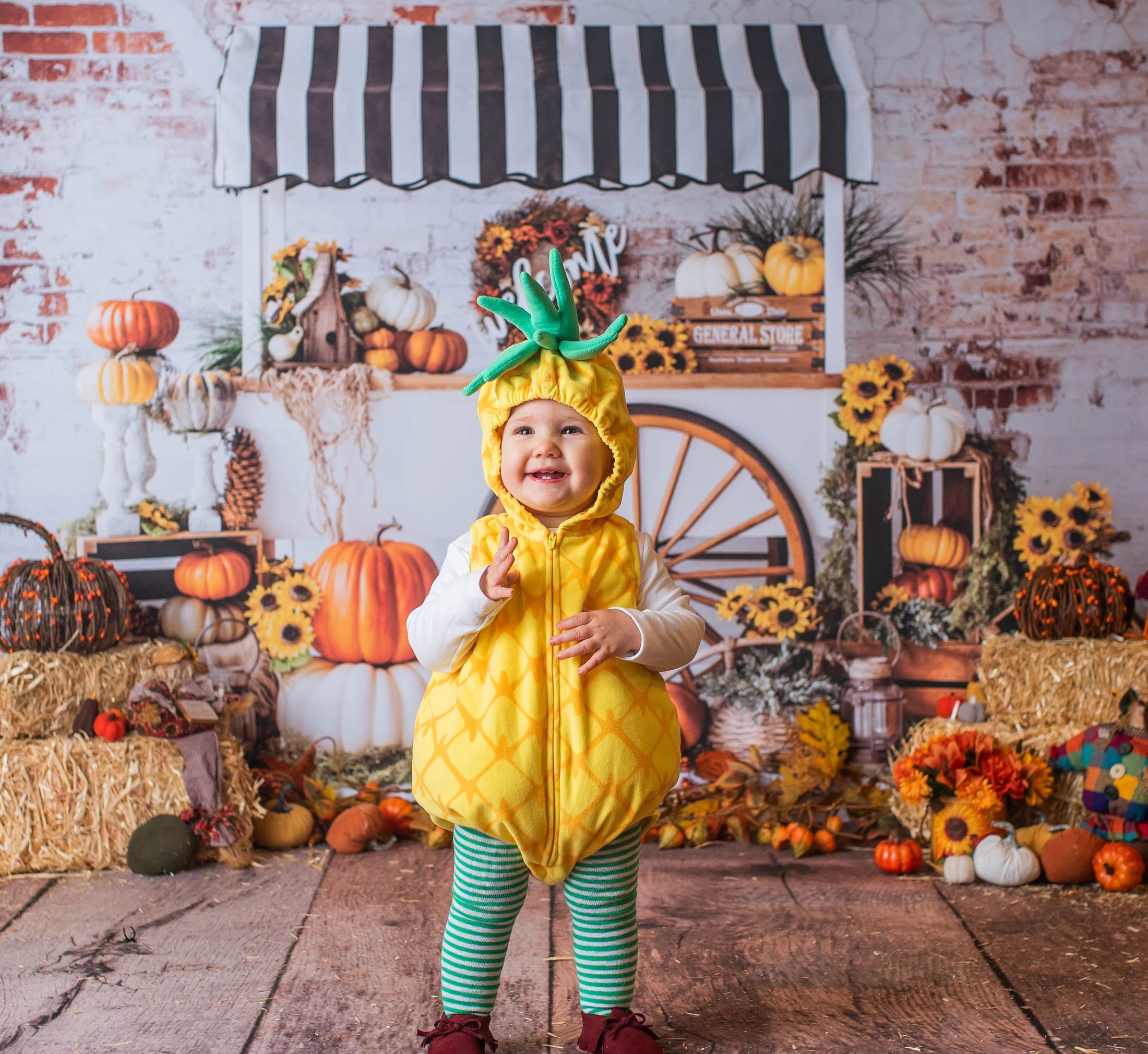 A happy toddler smiles while standing in a yellow pineapple costume. The background is a fall market scene with pumpkins, hay bales, and sunflowers.