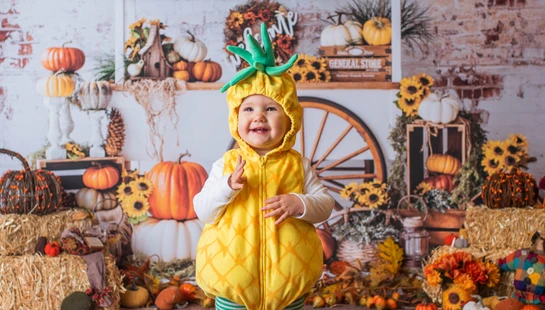 A happy toddler smiles while standing in a yellow pineapple costume. The background is a fall market scene with pumpkins, hay bales, and sunflowers.