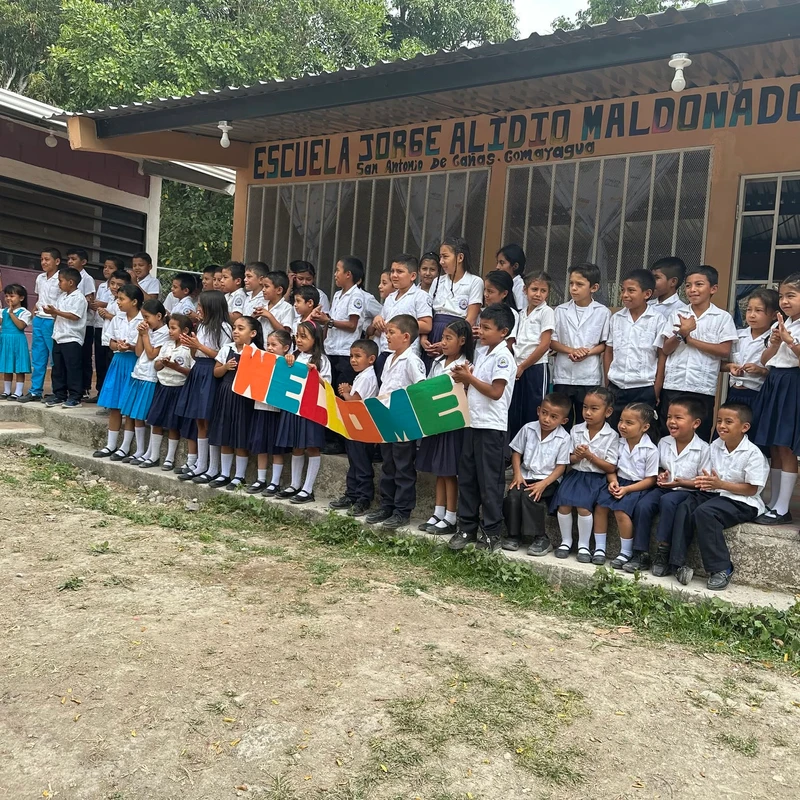 A large group of smiling schoolchildren in uniforms stand on the steps of their school, holding a colorful sign that spells out the word 'WELCOME'.