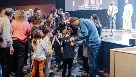 A group of adults and children stand together in a circle, bowing their heads in prayer in front of a stage during a church service.