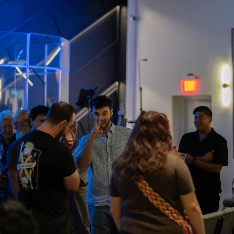 A man in a blue button-down shirt smiles and flashes a peace sign while standing and talking with a group of people indoors at an event.