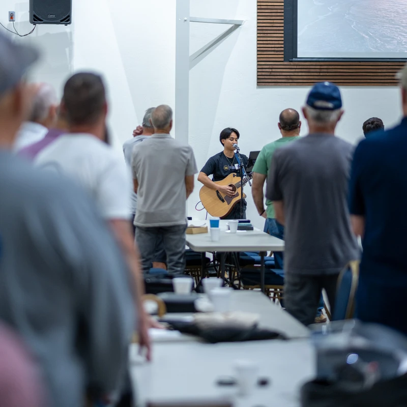 A young man with an acoustic guitar leads a group of standing men in worship. The camera is positioned behind the crowd, looking towards the performer.