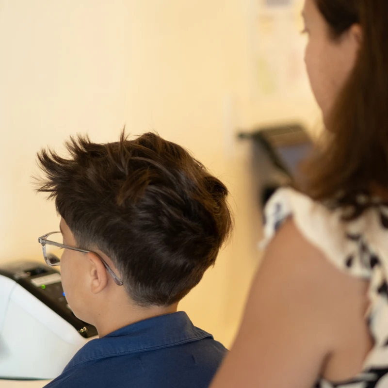 A young boy and his mother, seen from behind, use a wall-mounted tablet to check in. A classroom assignment chart is visible on the wall.