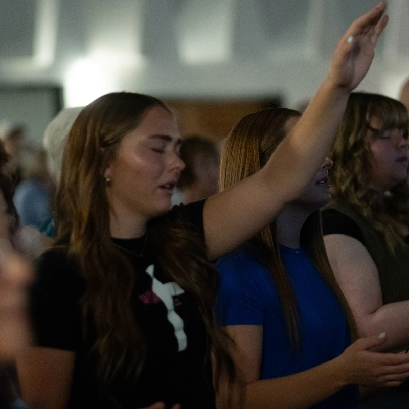 A group of women stand with their eyes closed in a dimly lit room. One woman in the center has her hand raised in a gesture of worship and prayer.