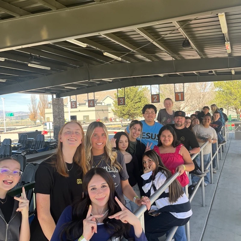 A large group of young people smiling and posing for a photo while waiting in a queue with metal railings at what appears to be a go-kart track.