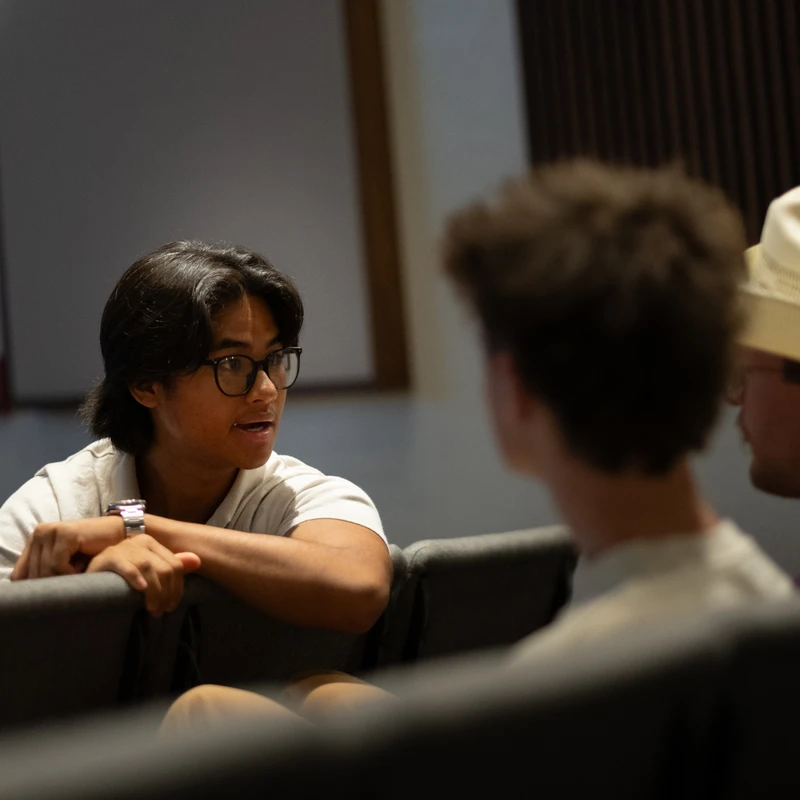 A young man with glasses turns in his seat to talk to two other young men sitting behind him in an auditorium. One wears a white cowboy hat.