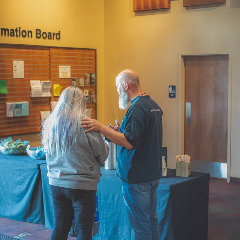 A bald man with a long white beard places a hand on a woman's shoulder as they stand and talk in a lobby next to a table with refreshments.