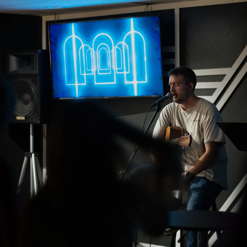 A man sits on a stage playing an acoustic guitar and singing into a microphone. Behind him is a screen with a glowing blue graphic in a dimly lit room.