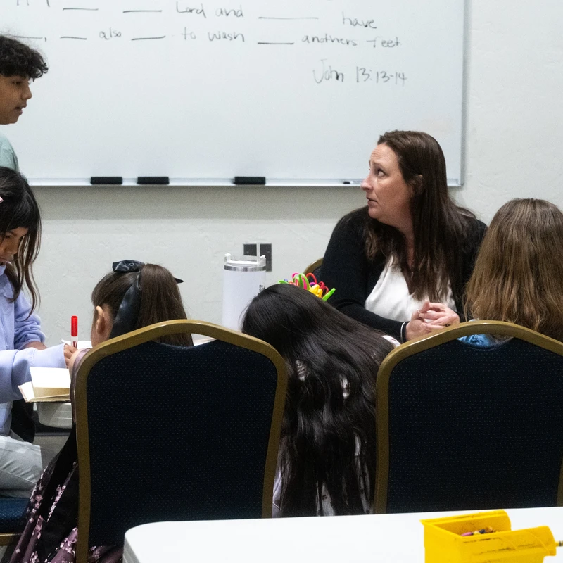 A teacher and several children are gathered around a table in a classroom, with a boy standing near a whiteboard with a bible verse written on it.