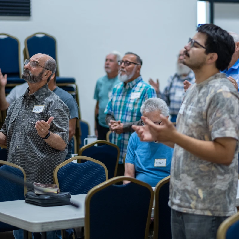 A group of men of various ages stand together in a room, with many lifting their hands and looking upwards in a gesture of prayer or worship.