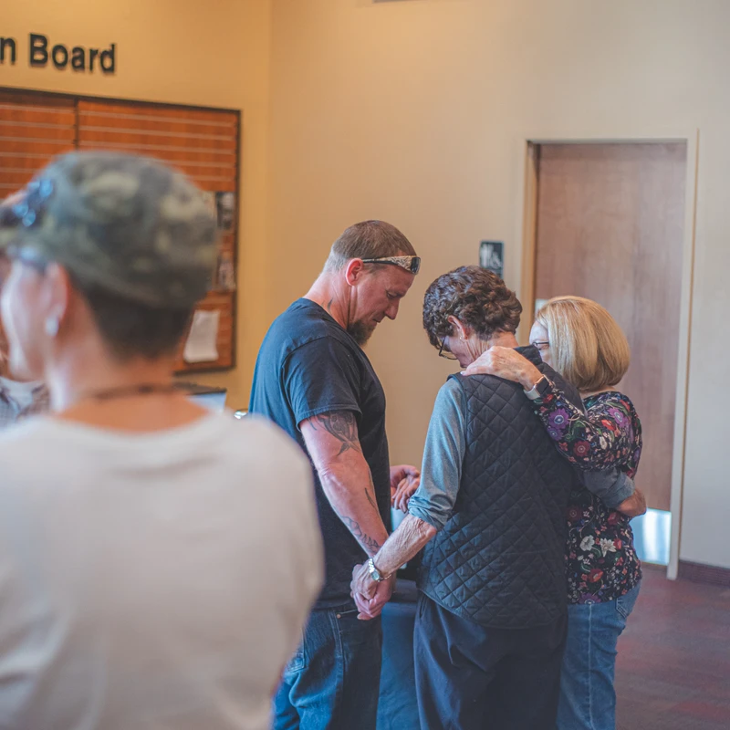 A man and two women stand in a circle, holding hands with heads bowed in prayer. Another woman embraces one of them from behind in a gesture of support.