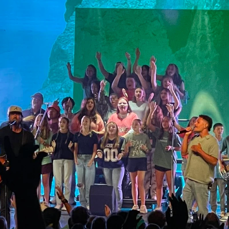 A band and a large youth choir perform on stage for an audience. Two male lead singers are at the front, with the choir singing enthusiastically behind them.