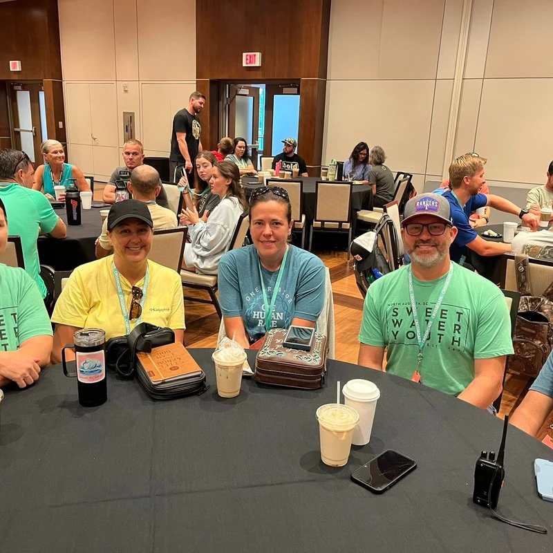 A group of five adults smiling for the camera while sitting at a round table with drinks in a large conference hall during an event.
