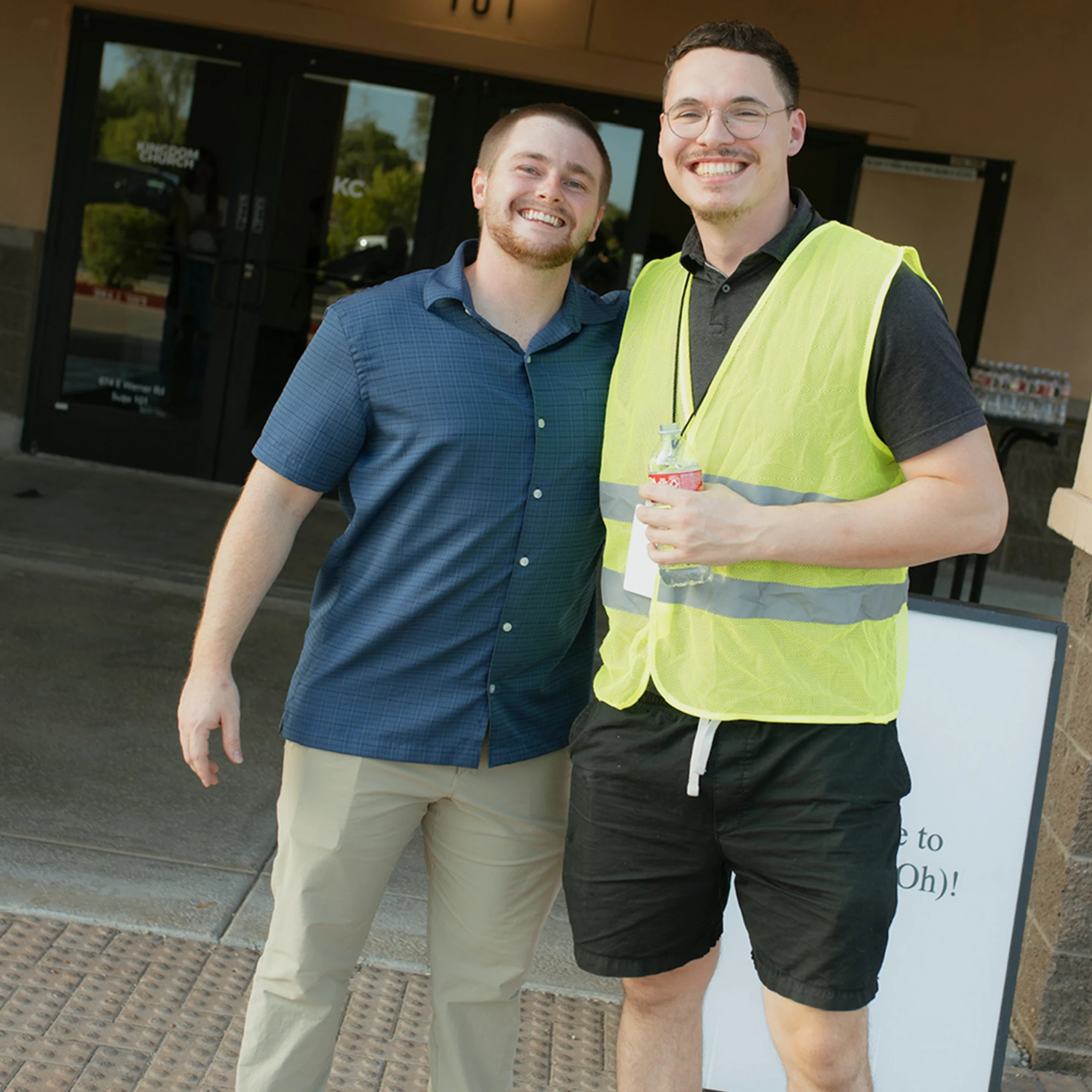 Two young men stand side-by-side and smile for the camera outside a building entrance. One wears a blue shirt, the other wears a yellow safety vest.