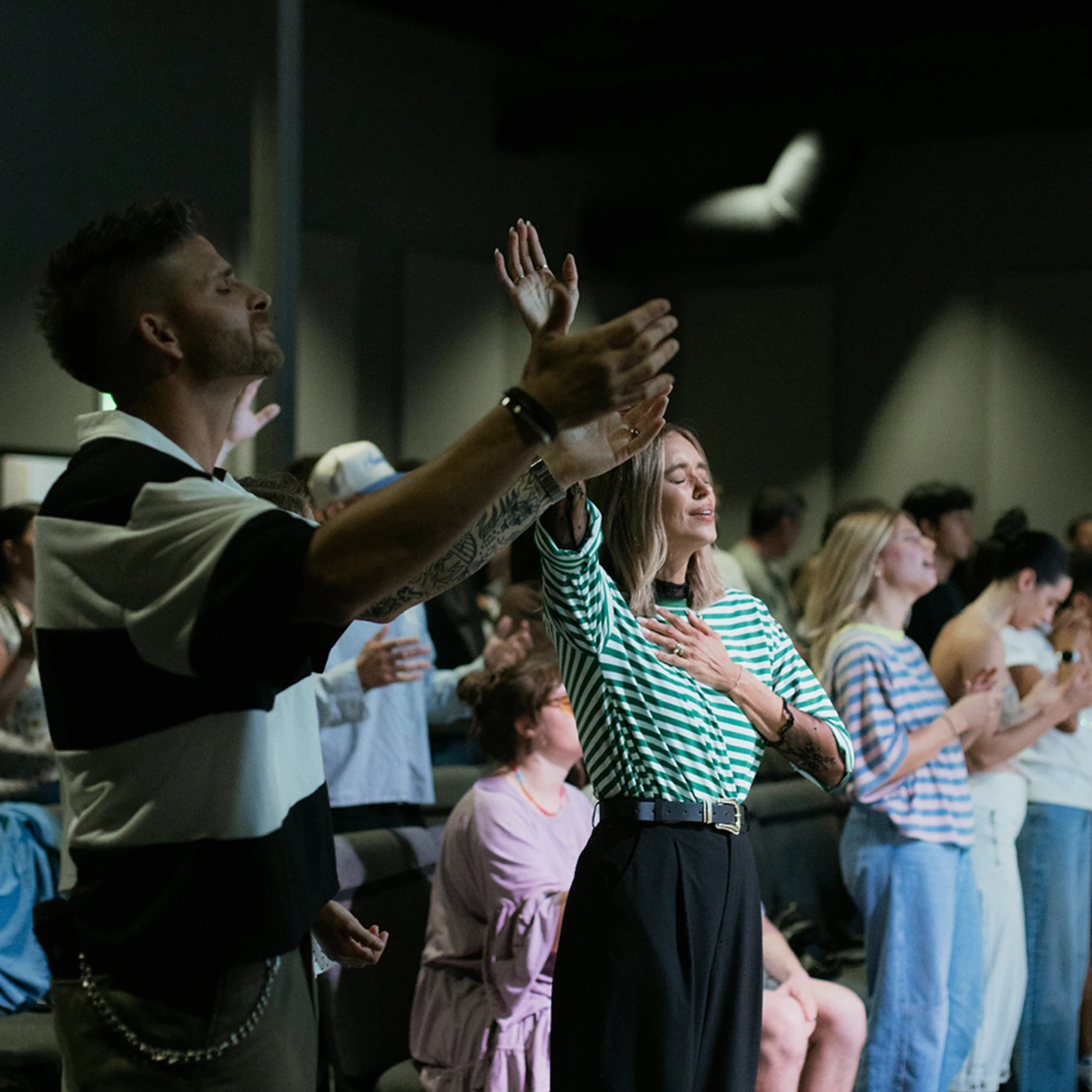 A diverse group of people stand with eyes closed and hands raised in worship during a church service. A man and woman are in the foreground.