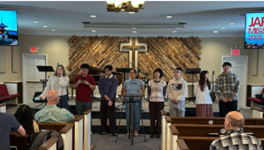 A group of young adults stands on a church stage, presenting to a congregation. Screens on the wall display information about a Japan missions trip in 2026.