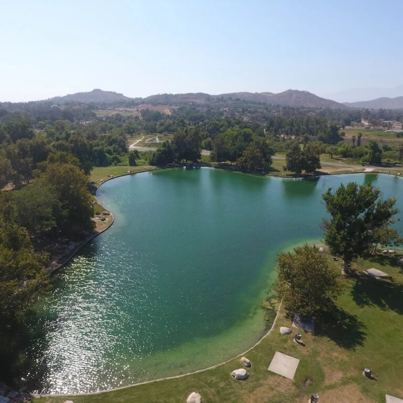 An aerial photo of a serene turquoise lake surrounded by green lawns and lush trees, with rolling hills visible in the hazy background under a clear sky.