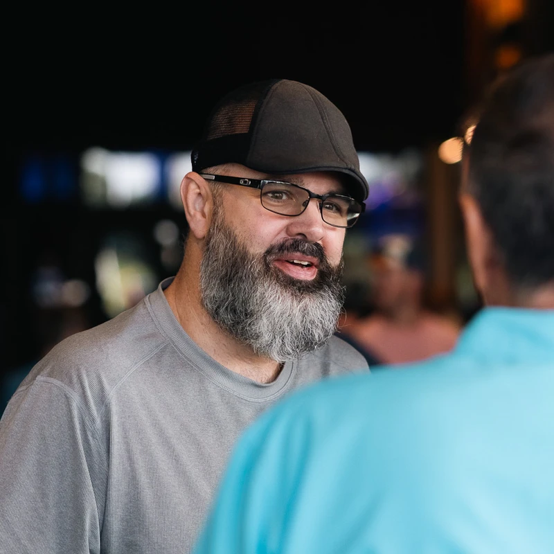 A man with a salt-and-pepper beard, glasses, and a dark flat cap talks to another person whose back is to the camera. He is wearing a gray shirt.