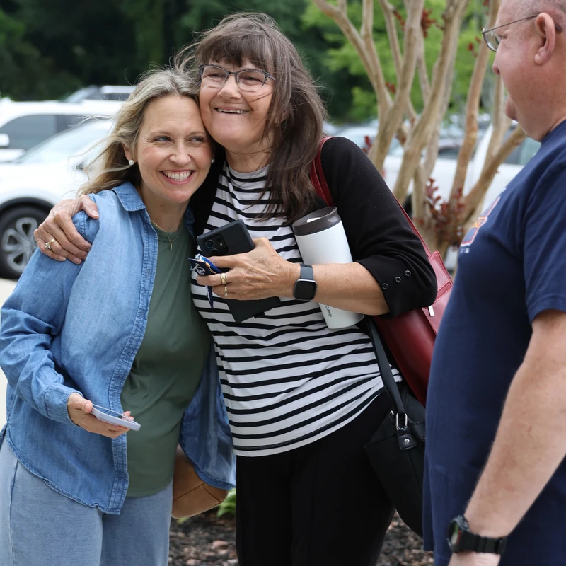 Two women smiling and hugging each other warmly outdoors. One wears a denim shirt and the other wears a striped shirt and glasses. A man is partially visible.