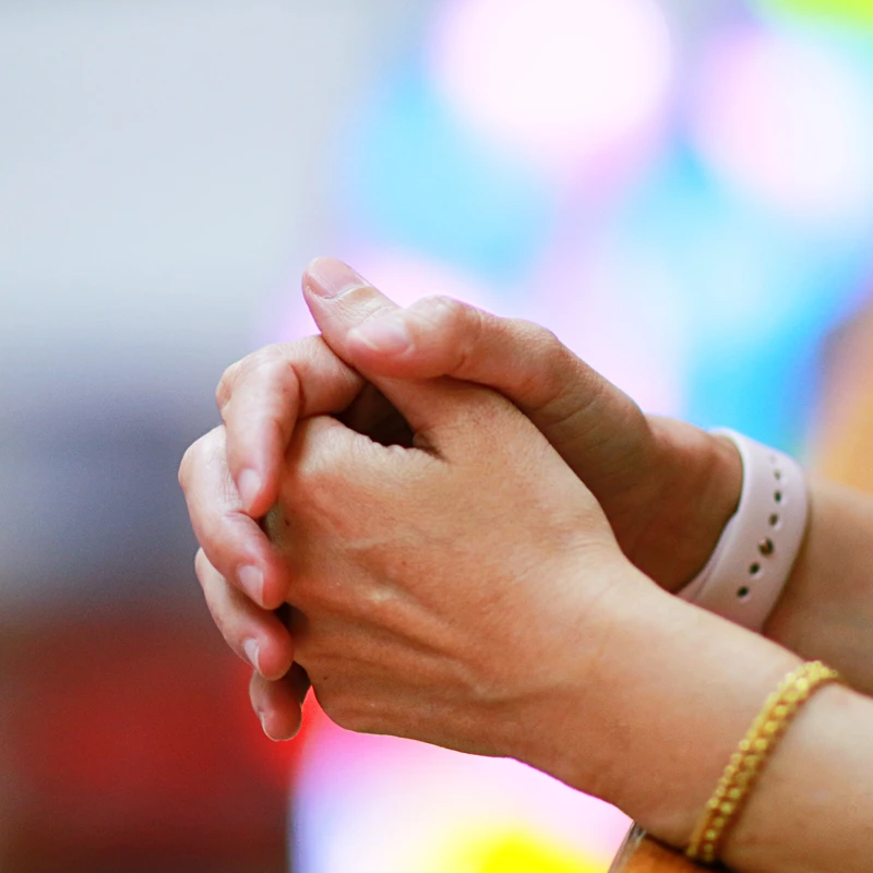 A close-up shot of a person's hands clasped together, wearing a watch and a bracelet, with a softly blurred, colorful background.